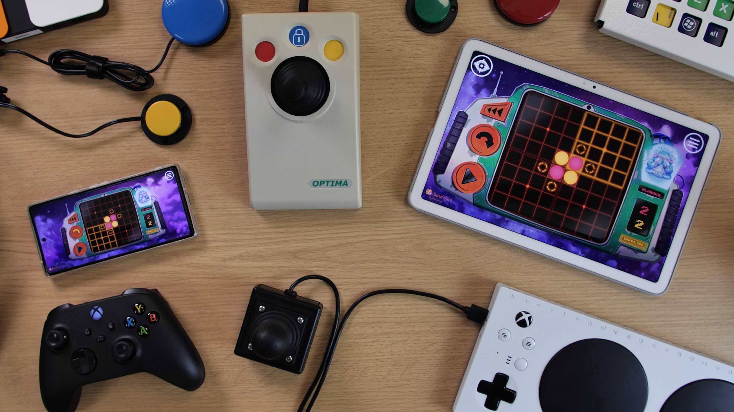 Photo showing an Android smartphone and a tablet with the game 'Othello' open on them. The devices are surrounded by devices including an Xbox Adaptive coController, and Optima Joystick, an Xbox controller and some accessibility switches.
