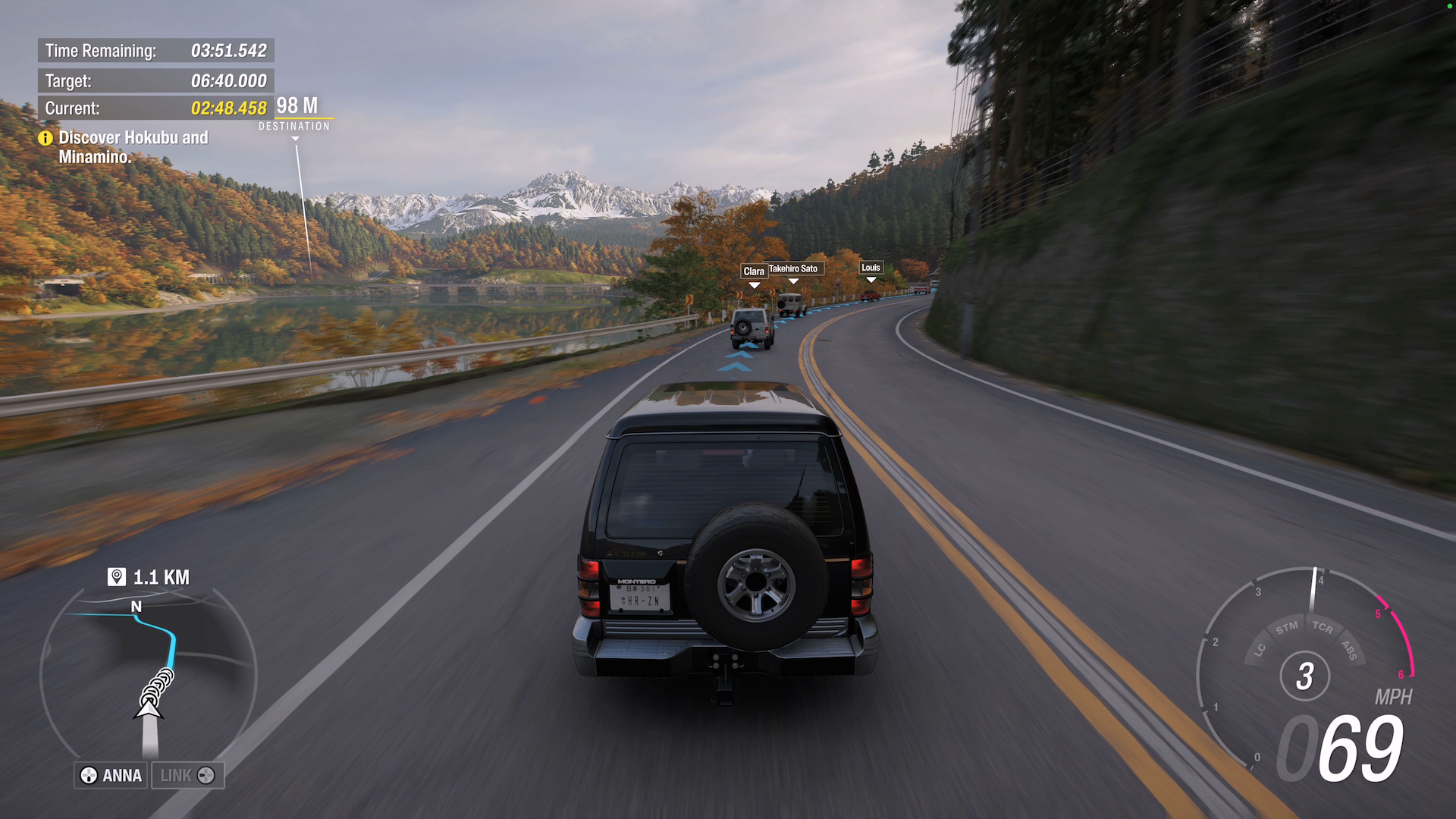 Screenshot showing a 4x4 vehicle driving along a road in the country with snow covered mountains in the distance and autumn coloured tress along the road and beyond.